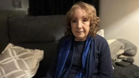 An older woman with pale curly hair sits on a grey sofa smiling.