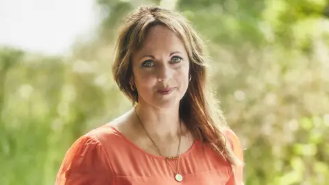 Josh Adam Jones Jenny Evans smiles at the camera. She is wearing a coral pink dress and a necklace. Her blonde hair is pulled to the side and is resting on her shoulder. Behind her is a blurred background of a garden.
