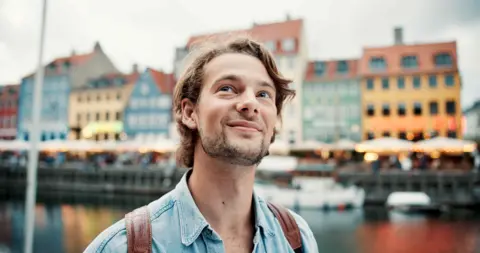 Getty Images A portrait of a happy wearing a denim shirt and a backpack. He has a happy smile as he looks around. There is a river and colourful buildings in the background. 