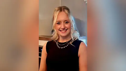 Family handout A young woman is wearing a sleeveless black top and a necklace, and has long blonde hair. She is smiling at the camera indoors.