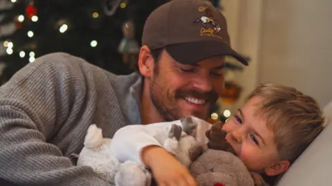 Ryan Libbey Ryan smiling and tickling his giggling young son Leo on the sofa