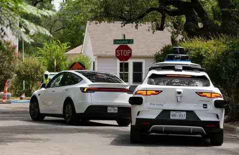 Austin American-Statesman via Getty Images  A driverless Tesla Robotaxi, left, and a Waymo autonomous vehicle make their way through road work area on a residential street in Austin, Tuesday, July 1, 2025.