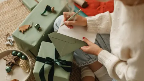 Getty Images A woman sitting on the floor with a pen in her right hand about to write a card to attach to a christmas present wrapped in green paper with a ribbon tied in a bow. She is surrounded by wooden Christmas decorations.