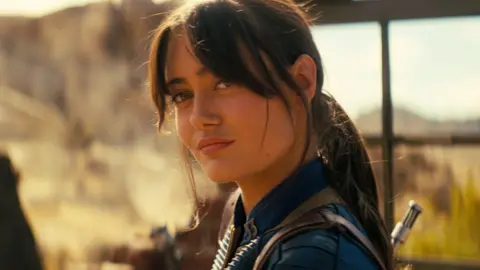 Prime Video A shot from Fallout shows Ella Purnell in her blue vault suit, hair tied back, looking quizzically at something in the distance. She's surrounded by desert and bathed in a dusky yellow sunlight. 