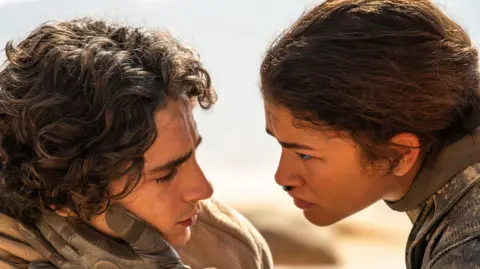 Legendary Pictures/Warner Bros. Zendaya lovingly holding Timothée Chalamet's head as they lie in the desert in Dune: Part Two