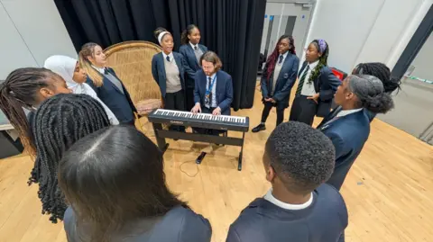 Dan Nelson / BBC A male music teacher sits at a piano in a school hall, surrounded by 12 Year 11 pupils in a circle around him. The pupils are singing. 