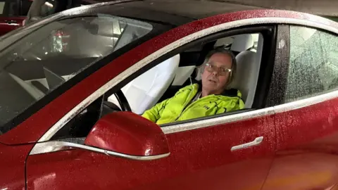 Ernest Gorrie Ernest Gorrie, in a neon yellow raincoat, sits in the drivers seat of a red Tesla