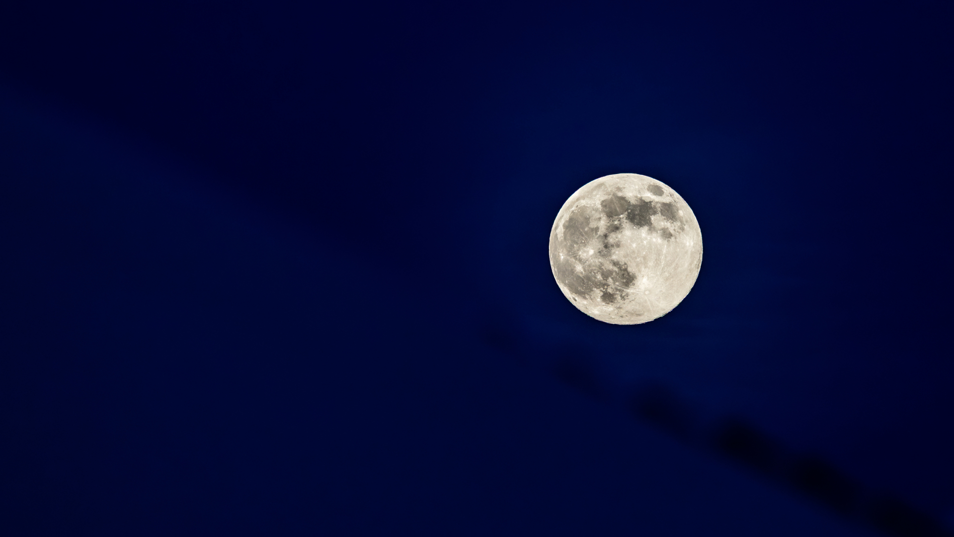 A full moon is pictured shining silver against a dark blue sky, as dark patches called lunar seas mark its surface.