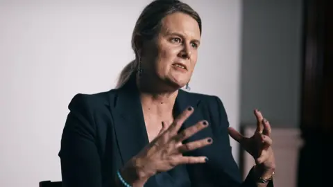 Alice Edwards is wearing a dark suit, and has brown hair tied back in a pony tale. 