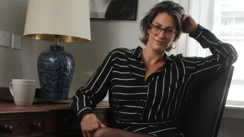 Image Angel Madelaine Thomas is sitting at a desk, turned towards the camera smiling. She is wearing a black and white striped shirt dress and sheer black tights. She has short dark hair with a blonde streak at the front and is wearing oversized reading glasses. On the desk there is a lamp with a blue base and a cream shade and a white mug. 