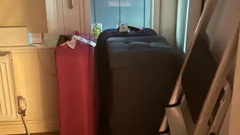 Masterson family Three large suitcase are pushed against a PVC and glass door, with a step ladder in front of them. On the door, a sticker warning of CCTV is visible.