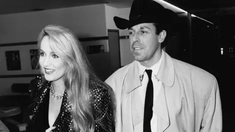 Getty Images A black and white photo of model Jerry Hall wearing a sparkly jacket sand fashion designer Antony Price wearing a black hat and trench coat in London in 1985.