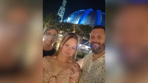 Family handout A young woman is standing in the middle of her mum and dad smiling at the camera.