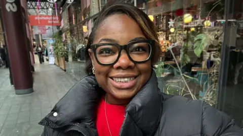 A selfie picture of Naomi, a 'Young Women's President' in the LDS in London. She is wearing a black puffer jacket and a red top underneath, and smiles while wearing glasses, stood on a shopping street.