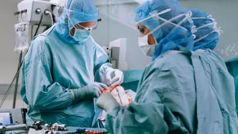 Getty Images A group of doctors wearing blue scrubs, masks and hairnets. They are in a surgical theatre performing a knee replacement operation on a patient. 