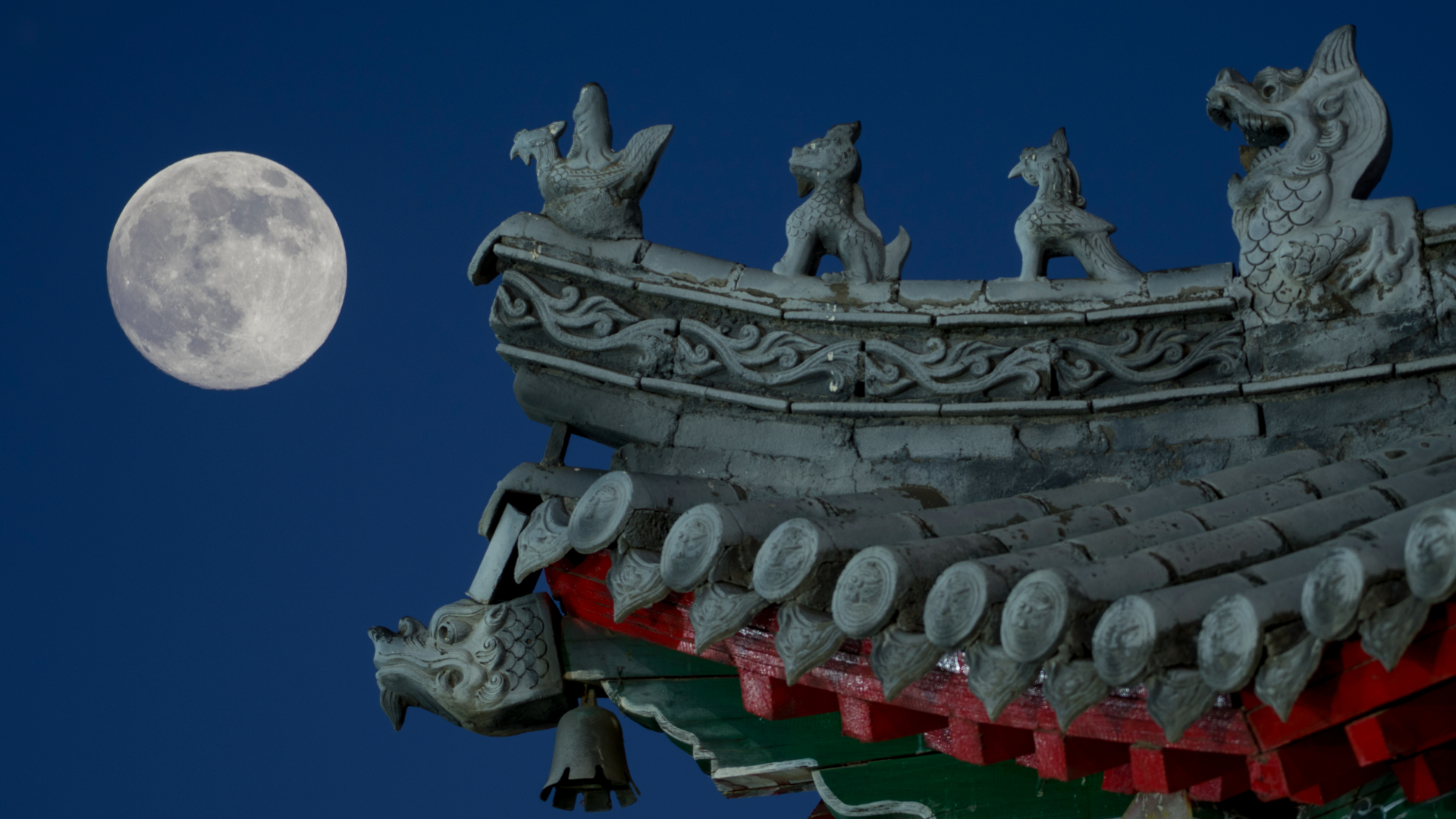A full moon is pictured glowing in a deep blue sky to the left of a traditional chinese carved stone roof.