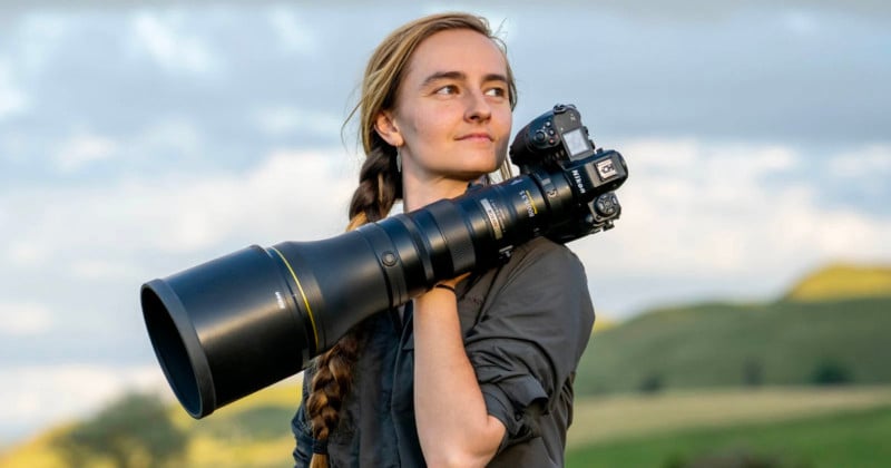 A woman with a long braid carries a large professional camera with a telephoto lens on her shoulder, standing outdoors with green hills and a cloudy sky in the background.