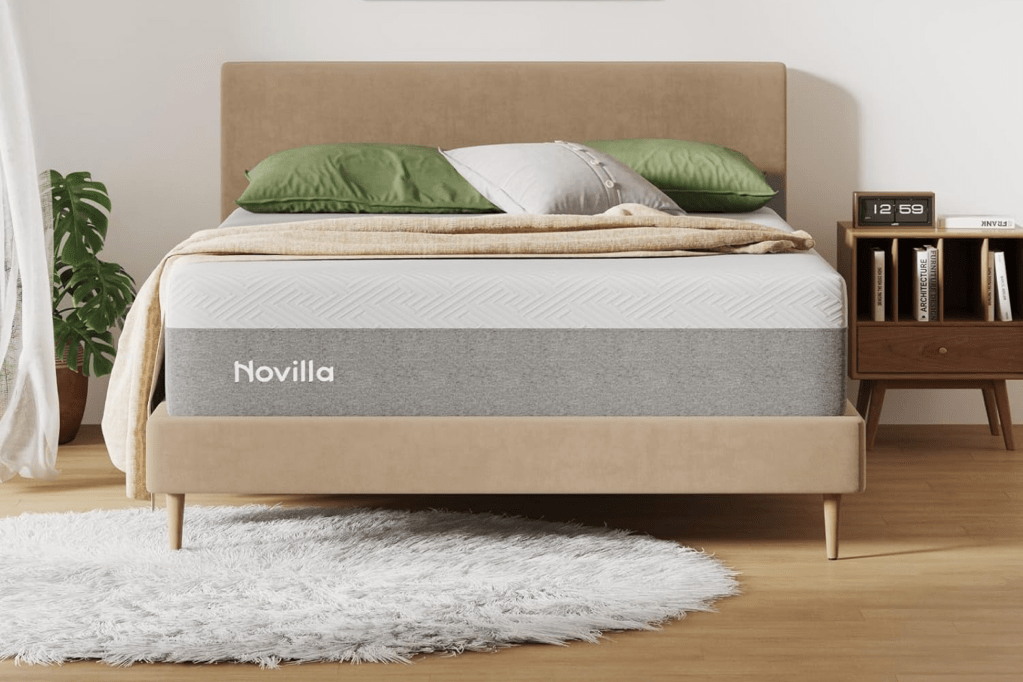 A Novilla mattress on a beige bed frame with a green and white pillow and beige blanket, next to a nightstand with a clock and books, and a plant.