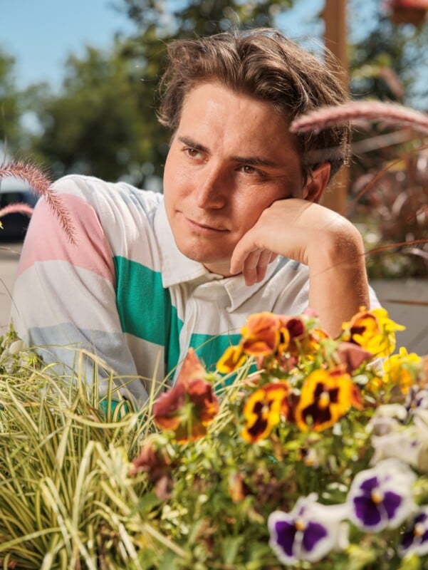 A young man with brown hair in a colorful striped shirt rests his chin on his hand and gazes thoughtfully at vibrant pansies and grasses in a sunny outdoor garden setting.