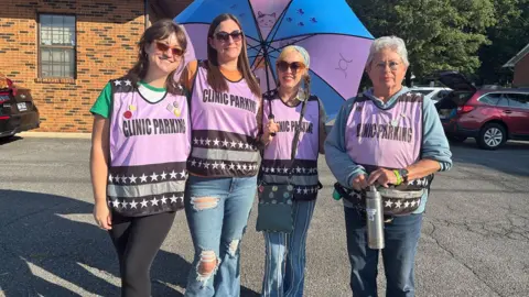 Four women stand outside the clinic in the parking lot, wearing pink vests that read "clinic parking". They all stand underneath a pink and blue umbrella to stay out of the sun