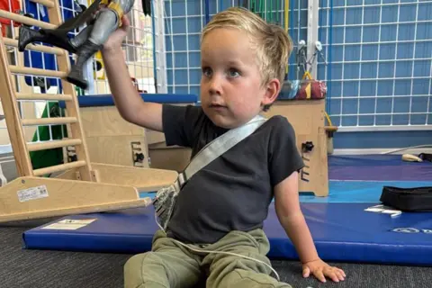 Contributed Edward is wearing a black T-shirt and green jogging bottoms and is sitting on the floor of a physiotherapy clinic and holding up a toy figure. 