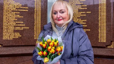 Jenni Hicks, with blonde hair, red lipstick and wearing a blue coat, stands before the Hillsborough memorial which has the names of the 97 engraved in golden lettering. She is holding a bunch of tulips.