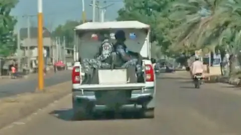 Gift Ufuoma/BBC A police 4x4 truck with two officers on the back and acting as a security escort drives along a tarmac road lined with trees in north-western Nigeria.