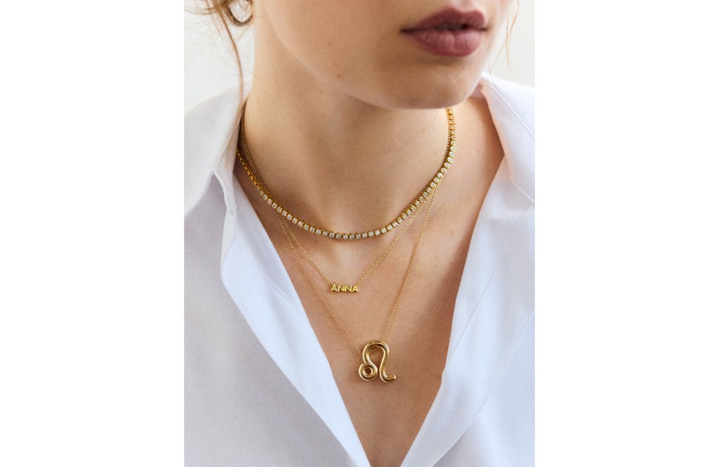 Three gold necklaces on a woman's neck, including a diamond tennis necklace, a "ANNA" nameplate necklace, and a Leo zodiac pendant.