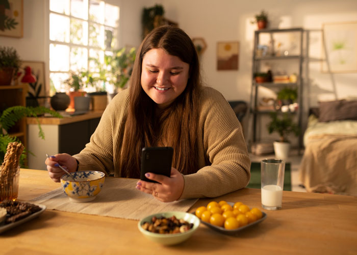 Young woman smiling at her phone while eating breakfast, reflecting on bro mocking sis over weight and diabetes. Young woman smiling at her phone while eating breakfast, reflecting on bro mocking sis over weight and diabetes.