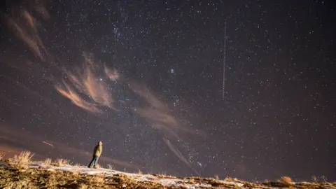 Getty Images A person stands on a snow-covered ridge at night looking at the sky, where a bright vertical Geminids meteor streak appears among numerous stars.