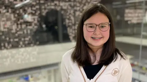 BBC A teenage girl with dark hair smiles at the camera, wearing a white cardigan