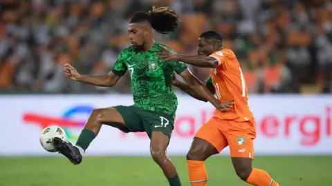 Getty Images Alex Iwobi kicks a football on a pitch. He wears a green patterned football jersey and has black dreadlocks. He is playing against Serge Aurier of Ivory Coast, who wears a bright orange football jersey. 