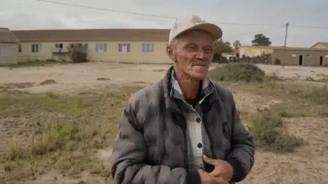 Andries Joseph shown from the waist up. He is wearing a grey padded coat and plaid shirt, plus a baseball cap on his head.