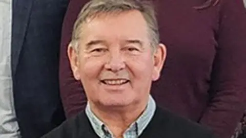 PA Peter Aitken wearing a blue collared shirt underneath a black jumper. He has grey hair and is smiling at the camera.