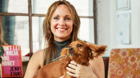 Josh Adam Jones Jenny Evans smiles at the camera and she is holding a brown puppy which has a pen in it's mouth. Behind Jenny is a book that says 'Don't let it break you, honey' by Jenny Evans. 