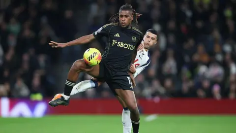 Getty Images Alex Iwobi kicks a football on a pitch. He wears a black Fulham jersey and has black dreadlocks. He is playing against Tottenham Hotspur's Pedro Porro, who wears a white jersey. 