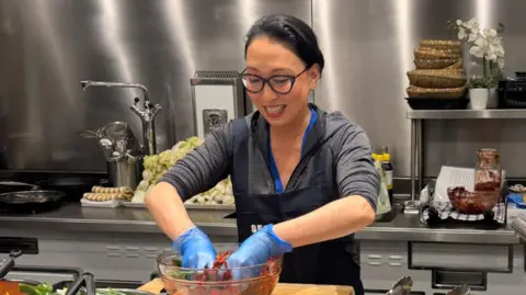 Judy Joo Korean-American TV chef Judy Joo teaching how to make kimchi from scratch in her central London flat. She is smiling at a kitchen counter and massaging red paste in a bowl with gloved hands