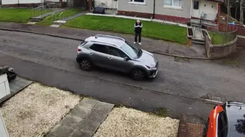 Masterson family A view of street - taken from CCTV on the first floor of a house - showing the side of a hatchback car mid-way through parking, while a white-haired man wearing dark trousers and a dark bodywarmer lights a cigarette on the kerb.
