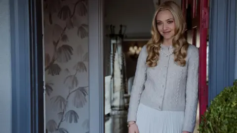 Lionsgate Actress Amanda Seyfried stands in a doorway wearing a grey knitted cardigan and white skirt with a pearl necklace. Her hair is blonde and curled.