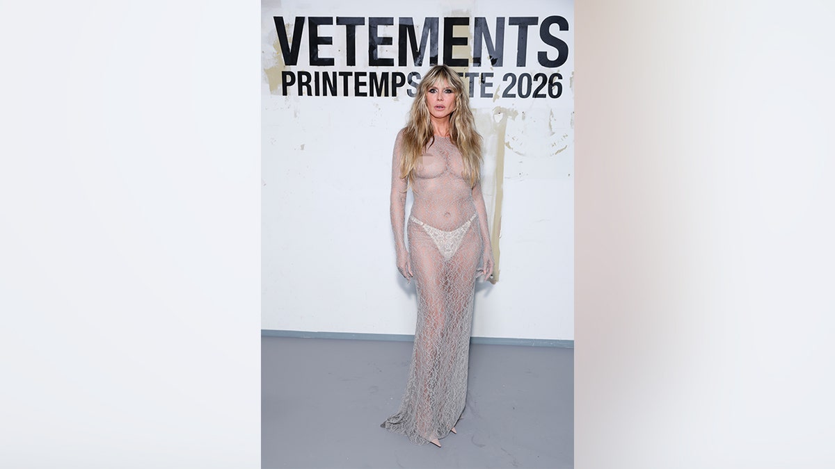 Heidi Klum stands against a Vetements backdrop at Paris Fashion Week wearing a floor-length nude lace gown with sheer fabric and visible underlayers.