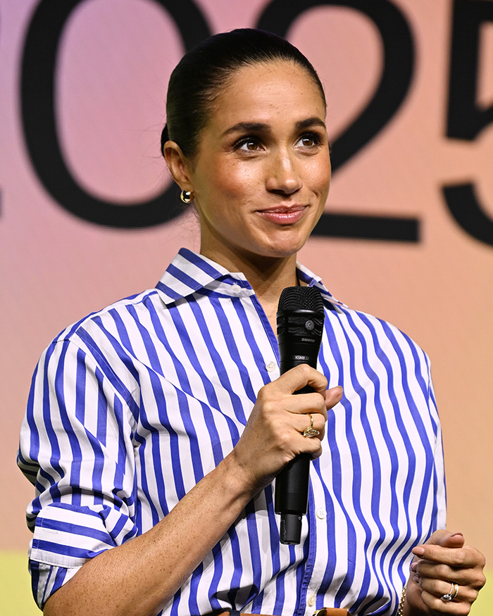 Meghan Markle speaking on stage holding a microphone with a striped blue and white shirt in an event setting. Meghan Markle speaking on stage holding a microphone with a striped blue and white shirt in an event setting.