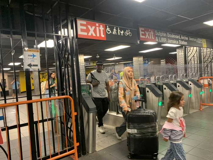 a line of people walk through an open gate in a Queens subway station