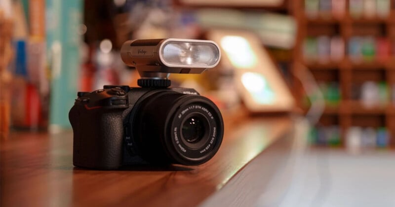 A digital camera with a large external microphone mounted on top sits on a wooden surface, with a softly blurred background featuring various colorful objects.