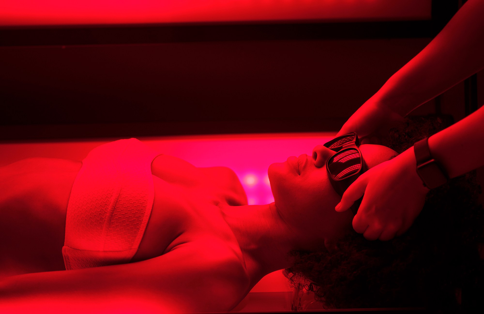 A person wearing eye protection lies under red light therapy, while hands massage their head.