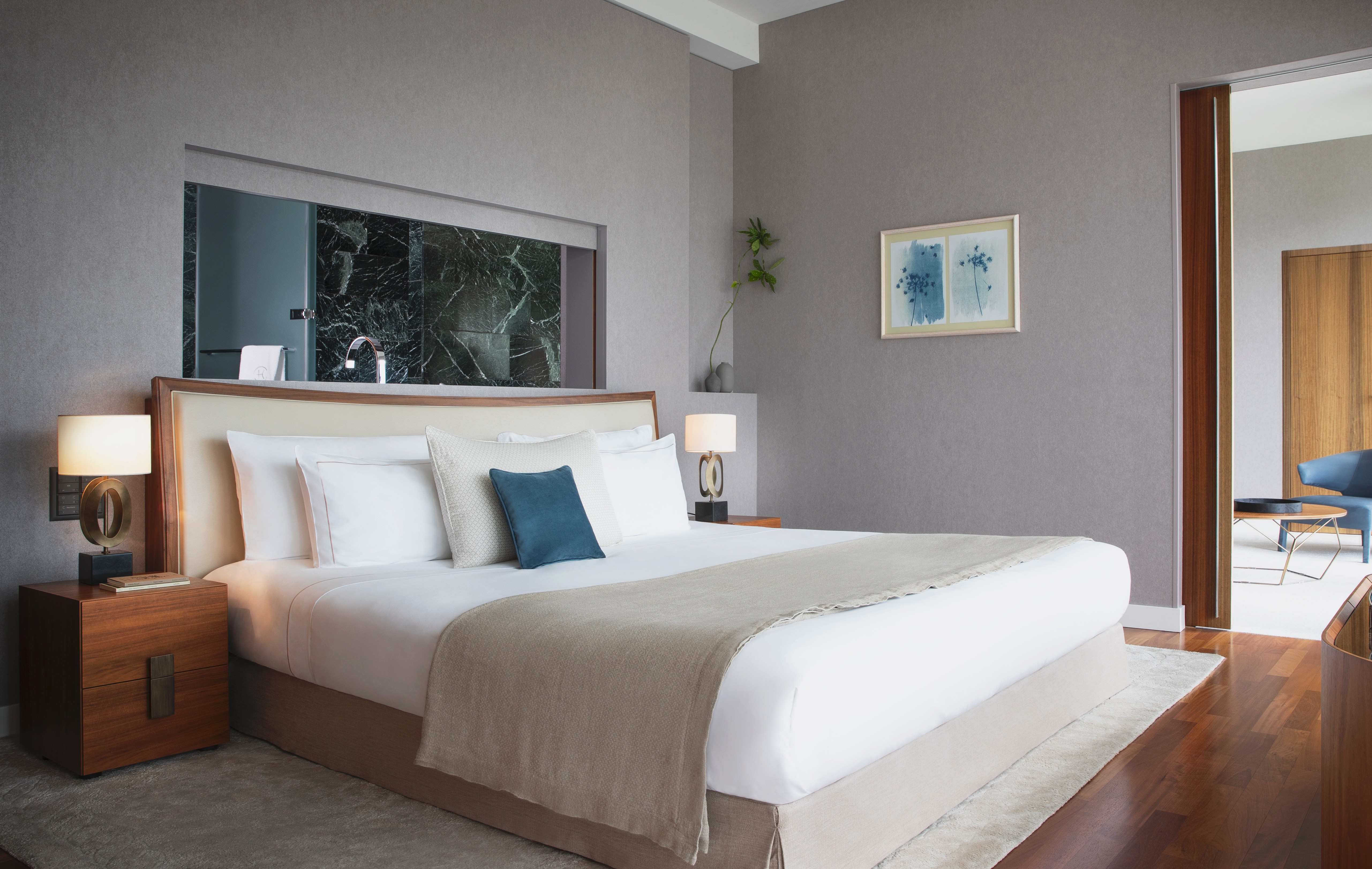 A modern bedroom with a large bed, two nightstands with lamps, a wall-mounted abstract picture, and an open doorway leading to another room.