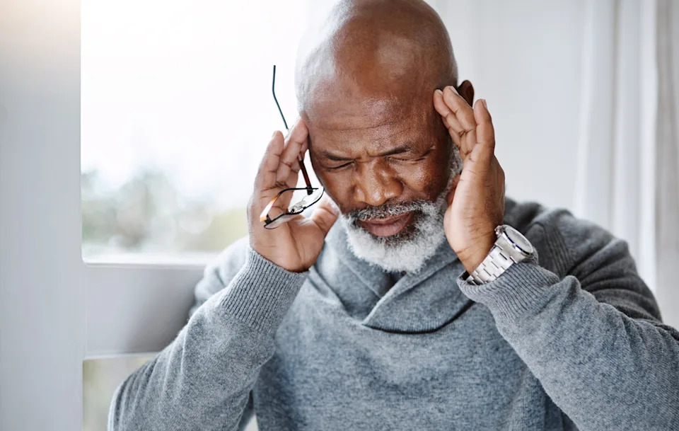 Man with glasses holds head, appearing stressed or in pain, wearing a cozy sweater near a window