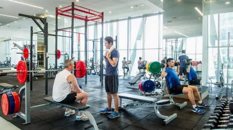 Getty Images People training in what looks like a commercial gym. One man is sat on a weight bench chatting to a man stood opposite him. In the right corner, a man is lifting dumbells with both arms. In the background, women can be seen using the bike machines. 
