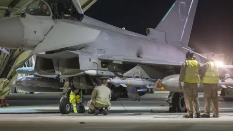 EPA/Shutterstock Royal Air Force ground crew preparing an RAF Typhoon fighter jet. It is night-time and the team are working under the floodlights of a hangar.