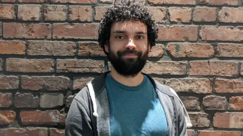Dr Nicholas Weise Nick Weise stands against a weathered brick wall. He has curly dark hair and a short beard, and is wearing a teal T‑shirt under a dark grey zip‑up hoodie. 