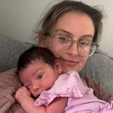 Chloe Chloe, a woman with light brown hair and clear rimmed glasses, sits with her daughter as a newborn baby lying on her and wearing a pink dress with frills.